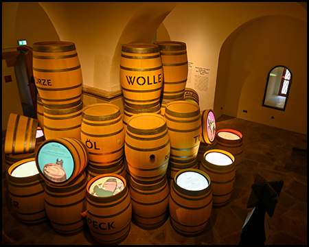Mehrere beschriftete Holzfässer, teils mit runden Bildschirmen versehen, in einem Ausstellungsraum.
