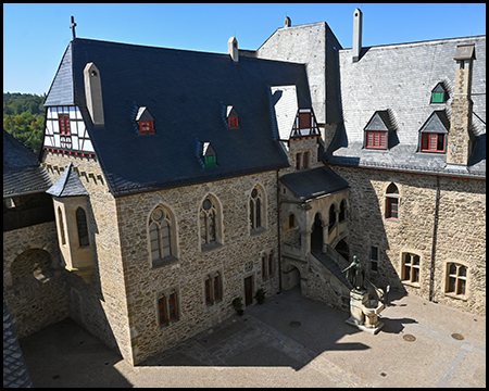Blick von oben auf einen Burginnenhof mit Fachwerkgiebel, Schieferdächern und einer Statue.