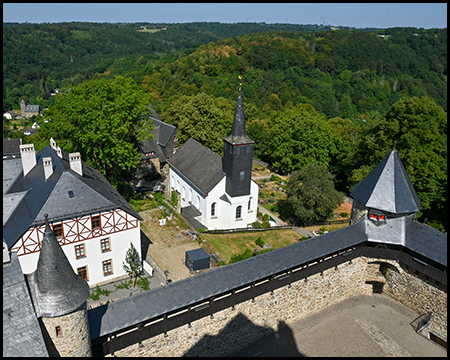 Blick von der Burg auf eine weiße Kirche mit Spitzturm, umgeben von Wald und Fachwerkhäusern.