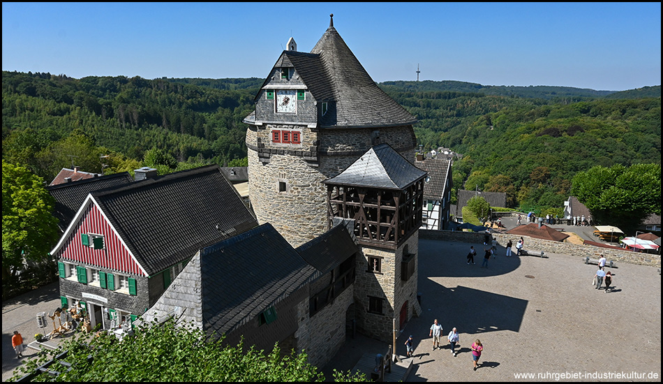 Blick von oben auf einen runden Burgturm mit Uhr und Glockenstuhl, umgeben von Wald und einem Platz mit Besuchern.