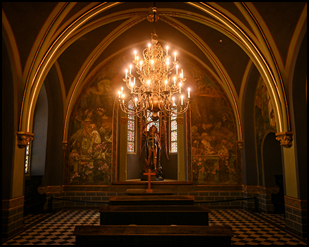 Eine Kapelle mit vergoldeten Gewölberippen, brennendem Kronleuchter und Wandgemälden im Altarraum.