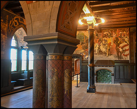 Ein Saal mit bemalten Säulen, Wandgemälden von Rittern und einem schmiedeeisernen Kronleuchter, Kamin im Hintergrund.