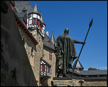 Eine bronzene Kriegerstatue mit Speer vor einem Fachwerkerker der Burg.