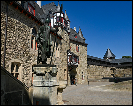 Eine Statue auf einem Sockel vor der Schlossfassade mit Fachwerkerker und Türmchen.