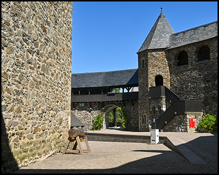 Ein Burghof mit Bruchsteinmauer, einem Turm und einem aufgeständerten Holzstamm