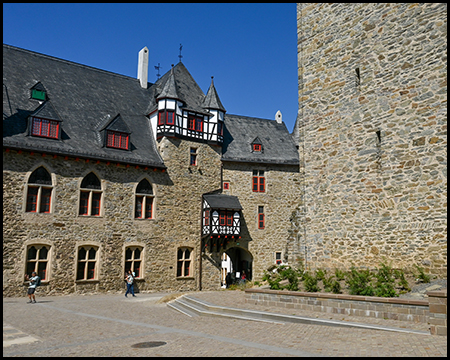 Ein Innenhof mit Fachwerkerker, roten Fensterrahmen und Besuchern vor einer hohen Steinmauer.
