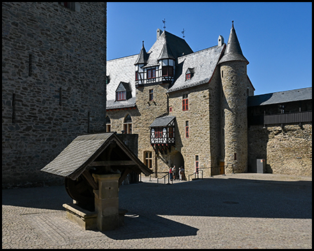 Ein hölzerner Brunnen vor der Schlossfassade mit Türmchen und Fachwerkerker.