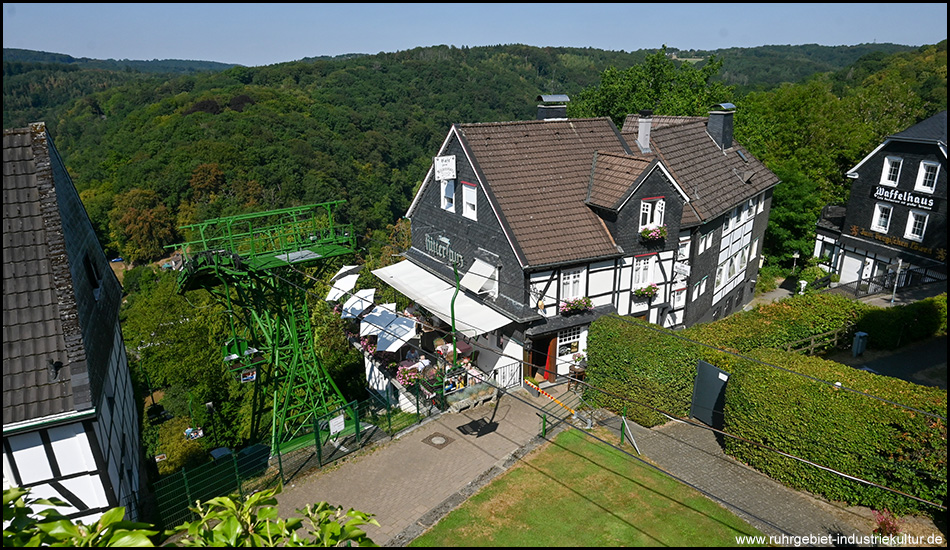 Blick von oben auf ein Fachwerkhaus mit Terrasse und die obere Station einer Seilbahn im Wald.