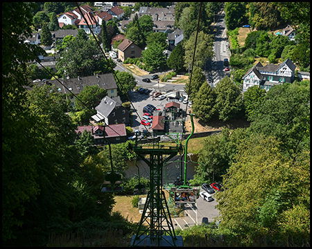 Blick aus einer Gondel auf einen Seilbahnmast und eine Talstation, umgeben von Wald und Häusern.