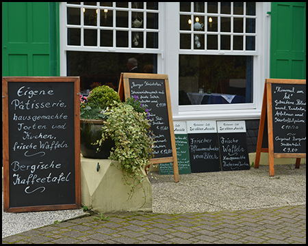 Mehrere Kreidetafeln mit handgeschriebenen Speisekarten vor einem Café mit grünen Fensterläden.