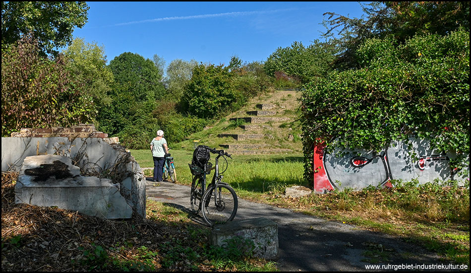 Ein grasbewachsener Hügel mit terrassenförmig angelegten Stufen und dunklen rechteckigen Öffnungen, die wie Eingänge oder Fenster aussehen. Ein Radfahrer mit weißer Kleidung steht links im Bild. Rechts sind Graffiti an einer Mauer und ein schwarzes Fahrrad zu sehen.