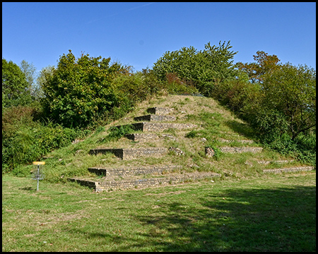 terrassenförmiger Grashügel in Form einer Pyramide, umgeben von grünen Bäumen und Sträuchern unter blauem Himmel.