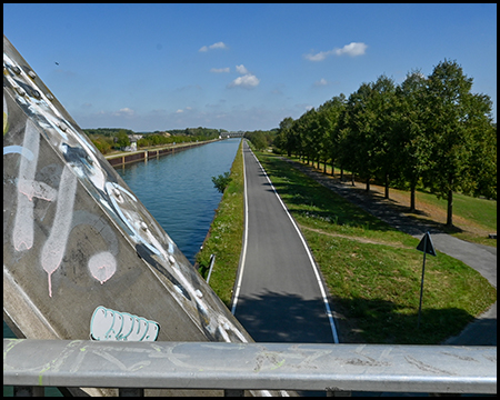 Blick auf einen schnurgeraden Kanal mit betoniertem Ufer. Links ist eine Brücke zu sehen, rechts führt ein Weg entlang des Wassers. Bäume säumen beide Seiten des Kanals unter einem blauen Himmel mit weißen Wolken.