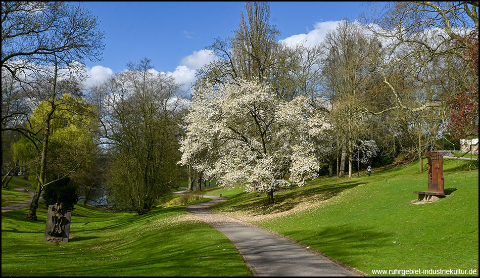 Parkanlage mit Wegen zwischen hügeliger Landschaft. Ein Baum fällt durch weiße Blüten auf. Einzelne rostige Stahlskulpturen stehen in der Landschaft