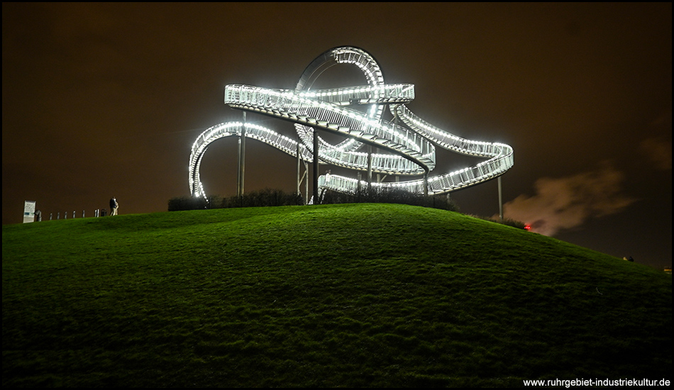 Beleuchtete Skulptur in Form einer Achterbahn auf einem nächtlichen Berggipfel