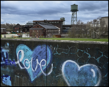 Industriegebäude und Wasserturm im Hintergrund. Im Vordergrund eine Mauer mit Grafitti mit Herzen darauf