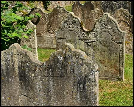 Mehrere verwitterte Grabsteine mit reliefartigen Verzierungen auf einem Friedhof.