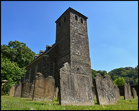 Ein quadratischer Kirchturm aus Bruchstein, davor eine Reihe alter Grabsteine.