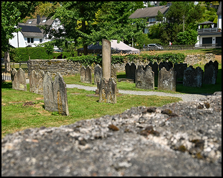 Reihen alter Grabsteine auf einem Friedhof, im Hintergrund Fachwerkhäuser.