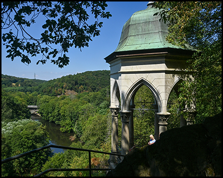 Ein steinerner Pavillon mit spitzbogigen Arkaden und grüner Kuppel auf einer Anhöhe, Blick auf ein bewaldetes Flusstal.