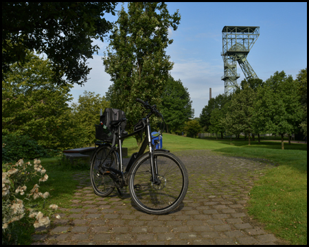 Ein Fahrrad parkt an einer Grünanlage. Im Hintergrund ein grünes Fördergerüst eines Bergwerks hinter Bäumen