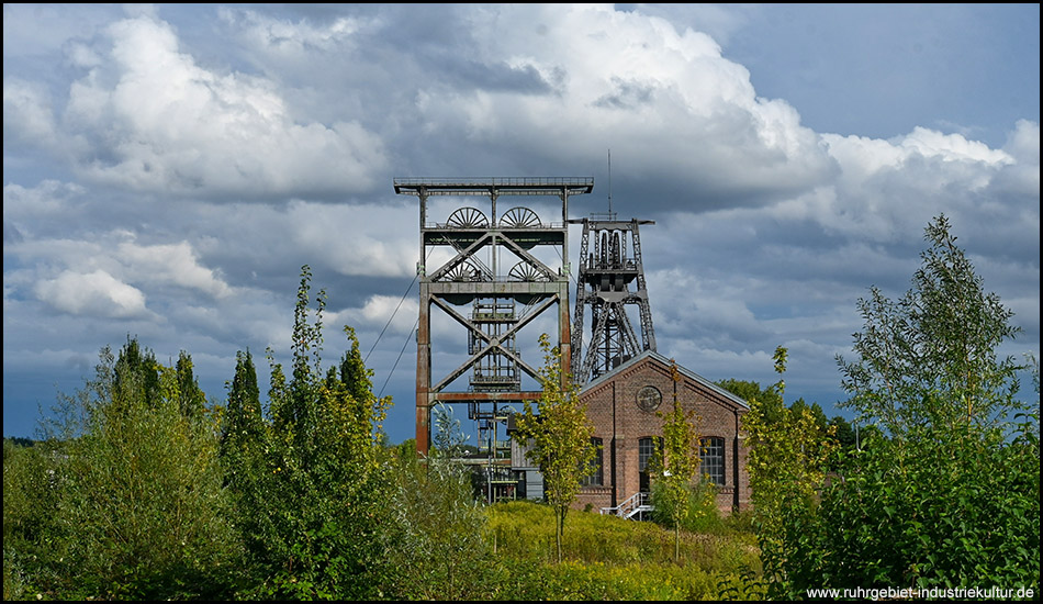 Historisches Zechengelände mit imposantem Doppelbock-Fördergerüst und Backsteingebäude, umgeben von grüner Vegetation unter dramatischem Wolkenhimmel